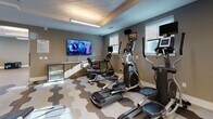 Located at 45 Coburg Rd in Charleston, SC, this single-floor fitness center is thoughtfully designed to accommodate a wide range of workout needs. The facility is divided into distinct functional zones, each with its own character and purpose. The main cardio and strength training area features a striking geometric hexagonal-patterned floor in neutral tones of gray, beige, and white, paired with a textured decorative ceiling that adds architectural interest. This zone is equipped with elliptical trainers, treadmills, and a multi-function cable machine, and includes a wall-mounted flat-screen TV and a built-in counter with a beverage cooler for convenience. Full-length mirrors expand the sense of space throughout. A dedicated free weights area provides a dumbbell rack, a weight bench, and additional cable resistance equipment, all set on the same distinctive hexagonal flooring. Separating these areas from the cycling studio are elegant sliding barn-style doors with frosted glass panels, offering both visual openness and acoustic separation. The cycling studio features smooth wood-look flooring, multiple spin bikes, a wall-mounted TV, and natural light streaming through several evenly spaced windows. The overall ambiance is clean, modern, and energizing, with cool gray-toned walls, bright overhead lighting, and a cohesive design aesthetic throughout.
