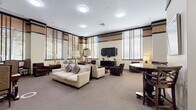 Located at 4720 Rosedale Ave in Bethesda, Maryland, this well-appointed community amenity space spans a single floor and offers a refined, welcoming atmosphere suited for both social gatherings and quiet retreats. The design is anchored by a generously sized lounge area featuring tall ceilings with decorative banded trim in warm tan and dark brown tones, providing a sophisticated architectural character throughout. A row of large windows dressed with wooden blinds lines the perimeter, flooding the space with abundant natural light while offering views of the surrounding greenery. The lounge is furnished with plush cream sofas, deep-toned upholstered armchairs, and dark wood accent pieces, all centered around a wall-mounted flat-screen television and a coordinating media console. Lush indoor plants add a natural, refreshing element to the room. A dedicated seating nook accommodates a round wooden dining table with ladder-back chairs, accented by framed artwork and table lamps, providing an intimate space for small meetings or casual conversation. An additional rectangular table with upholstered chairs near a set of double doors offers further flexible seating. The kitchenette is fitted with light wood cabinetry, granite-style countertops, a full-size refrigerator, and tile flooring, making it fully functional for gatherings and events.
