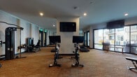 This fitness center at 1145 Chambers Rd in Columbus, Ohio, offers a comprehensive and well-appointed exercise facility designed to support a full range of workout routines. The single-level space is finished with durable textured carpeting throughout and benefits from recessed lighting that keeps the environment bright and functional.

The cardio section is prominently featured, housing a row of True-brand treadmills and elliptical machines, each equipped with individual screens for entertainment or workout tracking. Alongside these, dual stationary cycling bikes with digital displays provide dedicated options for low-impact training. A wall-mounted flat-screen television is positioned above the cycling area for shared viewing.

The strength training section features a selection of cable machines, a multi-function weight station, a leg press, and an adjustable bench, catering to both beginner and advanced users. A dedicated free weights area includes a rack stocked with medicine balls and kettlebells of varying sizes.

Full-length mirrors line multiple walls, visually expanding the space and allowing users to monitor their form. Large sliding glass doors on one side of the room flood the area with natural light and offer direct views of an outdoor courtyard. A ceiling fan overhead adds to the air circulation, and a small service station with storage cabinetry is available within the space.