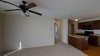This single-floor unit is designed with an open-concept layout that connects the living area and kitchen, creating a functional and cohesive space. The living area features plush wall-to-wall carpeting underfoot and neutral-toned walls that provide a clean, versatile backdrop. A ceiling fan with an integrated light fixture is centrally mounted, offering both comfort and ambient lighting. The space feels open and uncluttered, with smooth white ceilings contributing to an airy atmosphere.

The kitchen is positioned along one side of the space and features dark wood cabinetry that provides a warm contrast to the lighter tones throughout the unit. A raised breakfast bar with a built-in sink adds a practical counter space element, while overhead lighting keeps the area well-illuminated. Adjacent to the kitchen, a glimpse of additional storage or a pantry area can be seen, adding further utility to the layout.

A hallway visible from the main living area leads to additional rooms within the unit, hinting at a thoughtfully organized floor plan. The overall ambiance is neutral and move-in ready, offering a comfortable and practical living environment.