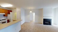 This single-floor space is designed with an open-concept layout that connects the kitchen and living areas seamlessly. The kitchen features a raised counter that serves as a natural divider, offering a pass-through view into the main living area. The living room is carpeted in a warm neutral tone and anchored by a fireplace with a classic black surround, adding a focal point to the space. A pendant light fixture hangs above, providing ambient illumination to the room's center. The walls are finished in a clean, bright white, contributing to an airy and open atmosphere throughout. A doorway visible from the living area leads further into the unit, suggesting additional rooms beyond. A mirrored surface near the kitchen entry reflects light and adds depth to the space. The overall layout is straightforward and functional, with consistent finishes throughout that give the space a cohesive, well-maintained appearance. The combination of neutral carpeting, white walls, and classic fixtures creates a versatile interior suitable for a variety of furnishing styles.