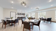 This thoughtfully designed single-floor communal space combines a bright, open dining area with a well-appointed kitchen and a private dining room, creating a versatile environment suited for both casual gatherings and formal occasions. The main dining area features light hardwood-style flooring throughout, with multiple seating arrangements — including square and rectangular tables paired with comfortable dark leather upholstered chairs. Decorative pendant and flush-mount light fixtures with cage-style designs add a contemporary touch overhead, while large windows along the perimeter flood the space with natural light and offer pleasant views of mature trees and landscaped grounds. A bar-height counter with pendant lighting and open shelving serves as a functional beverage and serving station. Adjacent to the main area, a fully equipped kitchen includes stainless steel appliances, rich dark cabinetry, a spacious central island with a white countertop, and additional built-in storage with floating shelves. A separate private dining room features elegant wainscoting wall paneling, a large conference-style table, gold wall sconces, and its own window view, offering an intimate and refined setting. The overall ambiance is clean, modern, and welcoming.