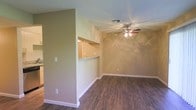 This single-story space presents a functional and straightforward layout with a cohesive design aesthetic throughout. The main living area features dark hardwood-style flooring that contrasts neatly with crisp white baseboards, lending a clean, grounded look to the room. The walls are finished in a warm tan tone, contributing to an inviting and neutral ambiance that suits a variety of furnishing styles.

A ceiling fan with a built-in light fixture is centrally positioned, providing both air circulation and ambient lighting to the living space. A standard HVAC vent is also visible in the ceiling, indicating a climate-controlled environment.

One of the more distinctive architectural features is a pass-through opening cut into the dividing wall between the living area and the kitchen. This pass-through is framed by what appears to be a granite or stone-topped counter ledge, offering a practical surface for serving or casual dining while maintaining a visual connection between the two spaces. The kitchen, partially visible through the opening, mirrors the warm wall tones seen throughout.

Overall, the space offers a simple, well-maintained interior with practical features and a warm, neutral palette.