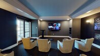 Located at 1554–1598 N Mill St in Naperville, IL, this single-floor amenity space is designed as a refined social lounge with distinct zones tailored for entertainment and relaxation. The layout flows across multiple connected areas, each with its own character while maintaining a cohesive, upscale aesthetic throughout.

The primary media lounge features deep navy and slate-gray walls beneath a tray ceiling with a contrasting blue inset, creating a cinematic atmosphere. Wall-mounted flat-screen televisions anchor the room, while plush cream swivel lounge chairs and low cocktail tables are arranged for comfortable group viewing. French doors allow access to the outdoors, introducing natural light to the otherwise moody interior.

An adjacent sitting area is distinguished by dark paneled walls adorned with framed black-and-white photography celebrating Chicago's architectural and cultural history, complemented by warm wall sconces that cast an inviting glow over the neutral lounge seating below.

A dedicated game room features a circular poker table surrounded by upholstered chairs beneath a coffered ceiling, with French doors offering views to the exterior. Textured wall treatments and recessed lighting give the space a relaxed yet polished feel.

A separate casual lounge area showcases exposed brick-style walls, a large wall-mounted television, leather club chairs, and a statement stone coffee table, with sliding glass door access to the outdoors. Throughout, patterned carpet flooring ties the interconnected spaces together cohesively.