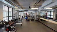 This fitness center offers a comprehensive workout environment spread across a single open floor. The space features an industrial aesthetic, with exposed ductwork running along the ceiling, concrete support columns, and durable dark rubber flooring throughout. Overhead fluorescent strip lighting ensures the facility is well-lit, complemented by natural light streaming in through large windows that provide views of the surrounding streetscape.

The cardio zone is generously stocked with treadmills, elliptical trainers, stationary bikes, and rowing machines, all arranged in orderly rows along the windowed perimeter. Mounted flat-screen televisions are positioned nearby for entertainment during workouts. A dedicated strength training area houses a variety of cable and weight machines, along with racks of dumbbells, kettlebells, medicine balls, and stability balls for functional fitness training. Full-length mirrors line one wall, supporting proper form and technique.

A free weights section provides barbells, weight plates, and adjustable benches, while a multipurpose open area offers space for stretching and bodyweight exercises. A wooden bench provides a rest area within the space. Artwork and motivational imagery on the walls add character to the facility, creating an engaging and energetic atmosphere suited for a wide range of fitness activities.