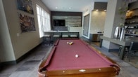 This single-floor recreational lounge is designed as a sophisticated entertainment hub. The centerpiece is a full-size pool table with a deep burgundy felt surface and classic wooden rails, positioned prominently within the open layout to allow ample room for gameplay on all sides.

The back wall features a striking design combination: a reclaimed wood-panel accent wall serves as a backdrop for a wall-mounted flat-screen television, flanked by a long console counter with decorative accessories. Adjacent to this, a textured white stone accent wall frames a dedicated wet bar area, complete with a beverage refrigerator and counter space — ideal for hosting guests.

Large windows along one side of the room flood the space with natural light, while recessed ceiling lighting provides consistent illumination throughout. Seating is thoughtfully integrated with upholstered stools positioned along a counter near the windows, offering a comfortable vantage point for spectators.

The flooring features a neutral-toned hard surface that complements the contemporary gray and charcoal color palette of the walls. Overall, the space strikes a balance between a modern, polished aesthetic and a relaxed, social atmosphere purpose-built for entertainment.