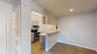 This single-floor residential unit offers a practical and modern layout designed for comfortable everyday living. The kitchen serves as a central highlight, featuring gray shaker-style cabinetry, white quartz countertops, and a full suite of stainless steel appliances including a refrigerator, electric range with over-the-range microwave, and a deep-set faucet at the sink. A peninsula breakfast bar with a clean white countertop separates the kitchen from the adjacent living area, creating an open and connected feel. Track pendant lighting illuminates the kitchen workspace, adding a contemporary touch. The living area is finished with durable wood-look flooring and opens directly to an outdoor patio through a full-width sliding glass door fitted with vertical blinds, allowing generous natural light to fill the space. A secondary room, carpeted and bright, features a ceiling fan with light fixture and a window with blinds overlooking a lush outdoor setting, making it well-suited as a bedroom. The unit's neutral color palette of whites and soft grays throughout creates a cohesive, clean aesthetic. Built-in shelving and ample storage options add to the unit's functionality, while white panel doors and crisp baseboards contribute to its polished, move-in-ready appeal.