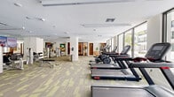 This well-appointed fitness center offers a generously sized, single-floor layout designed to accommodate a full range of workout activities. The space is defined by its contemporary aesthetic, featuring patterned green and grey carpeted flooring throughout and a clean white ceiling with integrated linear lighting that keeps the environment bright and energizing.

Along one side of the facility, a continuous bank of large, floor-to-ceiling windows floods the cardio zone with natural light while offering views of an outdoor pool area and surrounding greenery — providing an inspiring backdrop for treadmills, ellipticals, stair climbers, and stationary bikes lined up along the perimeter. A separate spin studio with glass-panel walls is accessible via double doors, offering dedicated cycling space.

The strength-training section is equally comprehensive, featuring a wide array of cable machines, multi-use weight stations, adjustable benches, and a dumbbell rack, all thoughtfully arranged to maximize usable floor space. Mirrors are strategically placed to enhance the sense of openness throughout the strength zone.

The facility also includes a small shelving unit and seating area, adding functional convenience. Vibrant artwork accents the walls, contributing a modern, polished atmosphere. Glass-paneled entry doors connect seamlessly to adjacent amenity spaces, reinforcing the facility's cohesive, upscale design.