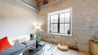 Located at 470 Market Ave SW in downtown Grand Rapids, MI, this one-bedroom, one-bathroom loft-style unit blends industrial character with contemporary design. The open living and kitchen area is anchored by striking exposed brick walls, warm wood beam ceilings, and visible industrial ductwork — hallmark features that give the space a distinctive urban loft aesthetic. Large windows flood the living area with natural light, enhancing the airy feel despite the unit's compact footprint.

The kitchen runs along one wall in a sleek galley configuration, featuring gray cabinetry extending to the ceiling, white subway tile backsplash, quartz countertops, and a full suite of stainless steel appliances. Light wood-tone flooring extends through the kitchen and living area, complementing the warm tones of the exposed ceiling above.

The bedroom is a cozy, well-defined space with a full-width sliding closet, offering efficient storage. The bathroom is cleanly finished with dark slate-style tile flooring, a white vanity with ample cabinetry and drawer storage, a large mirror with black-accented fixtures, and a separate tiled shower area. Each room reflects a cohesive modern design language, making efficient and stylish use of the available space throughout this character-rich urban unit.