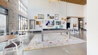 Located at 345 Great Circle Road in Nashville, Tennessee, this single-story community clubhouse delivers a dynamic blend of industrial and contemporary design. The space is anchored by dramatically high ceilings with warm wood slat detailing and bold sputnik-style chandeliers that serve as striking focal points throughout. Exposed brick walls lend an authentic, urban character, while large steel-framed windows and glass doors invite abundant natural light and views of the surrounding community.

The main gathering area is generously proportioned, featuring polished concrete floors and an open layout that accommodates multiple activity zones. A full-size pool table rests atop a colorful area rug surrounded by an eclectic gallery wall of artwork, while a wooden shuffleboard table offers additional recreation nearby. Comfortable lounge seating—including plush sofas and vintage-inspired chairs in muted greens and neutrals—creates inviting conversation areas alongside casual dining tables and modern wishbone-style chairs.

The lobby entry features a clean, functional design with a light wood credenza housing a self-serve coffee station, complemented by warm pendant wall sconces and a bold black city map mural. A curated gallery of Nashville-inspired artwork, including imagery of cowboy boots, guitars, and campfires, reinforces the local character throughout the space, creating a cohesive and energetic atmosphere.