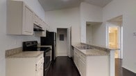 This single-floor residence offers a thoughtfully designed layout centered around a striking galley-style kitchen. The kitchen is a standout feature, boasting tall ceilings that amplify the sense of space and openness. White cabinetry lines both sides of the kitchen, providing generous storage, while light granite-style countertops offer ample workspace. Black stainless steel appliances, including an electric range and a full-size refrigerator, complement the clean, modern aesthetic. A double basin sink is conveniently positioned along the counter, and a dishwasher is seamlessly integrated into the cabinetry. Rich, dark hardwood flooring runs throughout the main area of the kitchen, contrasting beautifully with the white walls and bright overhead lighting. Light tile flooring can be seen transitioning into an adjacent room or hallway visible in the background, hinting at a well-connected floor plan. A door at the far end of the kitchen leads to additional areas of the home, suggesting a functional and flowing layout. The overall ambiance is clean, bright, and practical, making this space well-suited for everyday living and entertaining alike.