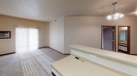 Located at 5170 Amber Valley Pkwy S in Fargo, North Dakota, this single-floor residential unit offers a practical and comfortable layout designed for everyday living. The open living area is finished with plush wall-to-wall carpeting and features a sliding glass door that allows natural light to fill the space while providing access to an outdoor area. A baseboard heating unit runs along the exterior wall, ensuring warmth during North Dakota's cold seasons. The kitchen is equipped with ample white cabinetry offering generous storage, light-colored countertops, a coil-burner electric range, and an over-the-range microwave. Tile flooring in the kitchen provides a durable, easy-to-clean surface. The bedroom features the same soft carpeting found throughout the unit, with neutral walls and multiple outlets, providing a versatile and restful retreat. The full bathroom includes a bathtub and shower combination, a vanity with storage, and tile flooring. A doorway from the bathroom leads directly into a carpeted walk-in closet outfitted with wire shelving, offering convenient and organized storage. Throughout the unit, warm wood-toned door frames and trim create a consistent and cohesive aesthetic against the neutral wall tones, contributing to an overall clean and understated ambiance.