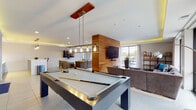 Located at 3443 Park St in Grove City, Ohio, this stylish single-floor community amenity space is designed to encourage socializing and recreation. The open-plan layout is anchored by a sleek billiards table with an industrial-style pendant light fixture suspended above, creating a natural focal point for the room. Warm wood accent walls add a rich contrast to the light gray walls and large-format tile flooring throughout, contributing to a contemporary yet inviting atmosphere. Ambient cove lighting traces the tray ceiling, casting a soft golden glow that enhances the modern aesthetic. Multiple distinct lounge zones are furnished with plush gray sofas and accent chairs, accented with pops of teal and yellow, and each oriented toward wall-mounted flat-screen TVs. A bar-height counter with industrial stools provides additional casual seating. Colorful curated artwork enlivens the walls near the reception and café-style area, which features pendant lighting and a workstation setup. Large sliding glass doors and floor-to-ceiling windows flood the space with natural light and offer direct views of and access to an outdoor deck area, seamlessly blending indoor and outdoor living. The overall ambiance is polished, energetic, and well-suited for both relaxation and social gatherings.