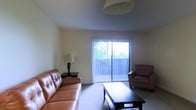 This single-floor space offers a clean and functional layout designed for comfortable everyday living. The living room serves as the central gathering area, featuring soft wall-to-wall carpeting underfoot and neutral-toned walls that create a calm, welcoming atmosphere. A large sliding glass door floods the room with natural light and provides direct access to an outdoor area, with vertical blinds offering adjustable light control and privacy. The room is furnished with a tufted leather sofa and a matching armchair, complemented by a rectangular wooden coffee table that anchors the seating arrangement. A classic table lamp on a side table adds a warm accent to the space. The ceiling is smooth and well-proportioned, with an overhead light fixture providing additional illumination. The overall ambiance is understated and practical, with a straightforward design that emphasizes comfort and usability. The connection between the interior and the lush greenery visible through the sliding glass door gives the living area a pleasant sense of openness, making it feel bright and airy throughout the day.