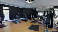 This fitness center offers a focused and functional workout environment designed to accommodate a variety of exercise routines. The space features vaulted ceilings with crown molding and a ceiling fan that provides airflow and adds to the room's comfortable atmosphere. Bold, dark-painted walls create a motivating, gym-like ambiance, contrasted by clean white trim and warm hardwood-style flooring. A large floor-to-ceiling mirror spans one wall, reflecting natural light from a window and visually expanding the space. The equipment layout is thoughtfully arranged, featuring cardio machines including a stationary bike, elliptical, and treadmill, as well as a multi-use strength training station. Protective rubber mats are placed beneath each piece of equipment to safeguard the flooring. A wall-mounted television is positioned for convenient viewing during workouts. The combination of practical design, ample equipment, and a clean, polished finish makes this fitness center a well-rounded facility suited for both cardio and strength training needs.