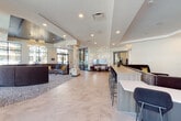 This contemporary single-floor clubhouse space is thoughtfully designed to foster both social interaction and relaxed leisure. The open-plan layout is anchored by large-format herringbone-patterned tile flooring that flows seamlessly throughout, lending a clean, upscale aesthetic to the entire space.

The main lounge area features a generous curved sectional sofa accented with bold navy and gold throw pillows, complemented by stylish ottomans and pendant lighting. A striking decorative column of natural birch trunks serves as a sculptural focal point, adding an organic contrast to the otherwise sleek, modern interior. Adjacent to the lounge is a well-appointed bar-style counter with bar seating, display monitors, and a kitchenette with a sink and cabinetry—ideal for casual gatherings.

Floor-to-ceiling windows and glass doors flood the space with abundant natural light and offer open views of the surrounding outdoor areas and courtyard. A dedicated game room provides an entertainment hub complete with a pool table, shuffleboard, arcade machines, and oversized Connect Four, all set against a boldly patterned area rug and atmospheric pendant lighting.

Throughout the space, curated artwork, textured accent walls, and recessed lighting contribute to a polished, welcoming ambiance designed for community connection.