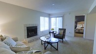 This single-floor residence offers a comfortable and well-appointed living environment defined by clean lines and a neutral color palette. The living room serves as the heart of the space, highlighted by a classic tile-surround fireplace that anchors the room and adds a cozy focal point. Large windows, dressed with vertical blinds, flood the area with natural light and offer views of the surrounding greenery, creating a bright and welcoming atmosphere. The soft, wall-to-wall carpeting complements the light gray walls, lending a warm and understated elegance to the interior. A textured ceiling adds subtle architectural character overhead. The open layout of the living area provides ample room for comfortable seating arrangements and decorative accents, making it well-suited for both relaxing and entertaining. The overall ambiance of the space is calm and inviting, with a cohesive design sensibility that balances simplicity with functionality. The combination of natural light, the fireplace feature, and the generous proportions of the living area come together to create a residence that feels both practical and welcoming.