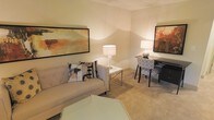 This single-floor space presents a well-appointed living area that balances comfort and functionality. The room is finished with soft, neutral-toned carpeting that adds warmth underfoot and complements the light-colored walls, creating a calm and welcoming atmosphere throughout.

The living area is furnished with a tufted light gray sofa accented by decorative throw pillows in abstract patterns, positioned alongside a sleek chrome side table with a contemporary lamp that provides warm ambient lighting. Large framed abstract artwork in warm earth tones—rust, amber, and brown—adorns the walls, adding visual depth and a modern aesthetic to the space.

A dedicated workspace is seamlessly integrated into the room, featuring a dark-framed desk and a patterned upholstered chair, making the space versatile for both leisure and productivity. A second accent lamp on the desk mirrors the warm lighting scheme, ensuring the entire room feels cohesive and evenly lit.

A glass-topped coffee table anchors the seating area, reflecting light and contributing to an open, airy feel despite the room's intimate scale. The overall ambiance is cozy yet polished, with thoughtful décor choices that make the space feel both functional and stylish.