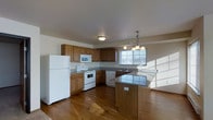 This single-floor space offers a practical and inviting layout centered around a thoughtfully designed kitchen. The kitchen is a standout feature, showcasing warm honey-toned wood cabinetry that provides ample storage and a classic, timeless aesthetic. Countertops finished in a granite-style surface add a touch of durability and visual interest, complementing the rich hardwood flooring that runs throughout the space.

A central kitchen island extends the counter space and serves as a natural gathering point, ideal for meal preparation or casual dining. The full suite of white appliances — including a refrigerator, electric range, built-in microwave, and dishwasher — is seamlessly integrated into the cabinetry for a cohesive look.

Natural light plays a significant role in the ambiance of this space. Large windows allow generous daylight to pour in, creating a bright and airy atmosphere. An elegant chandelier above the island area adds a decorative focal point and provides warm overhead lighting to complement the natural light.

The open layout connects the kitchen to the adjacent living area, promoting a sense of flow and spaciousness. The overall design balances functionality with comfort, making this a practical and welcoming environment suited for everyday living.
