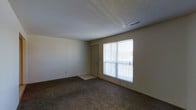 This single-floor space features a generously sized main living area finished with wall-to-wall carpet in a neutral tone, providing a comfortable and understated foundation. The room is illuminated by natural light streaming through a large window fitted with horizontal blinds, which overlooks what appears to be a balcony or patio, accessible via an adjacent exterior door. This combination of window and door enhances the sense of openness and connection to the outdoors. The walls are painted in a soft, neutral beige, complementing the overall light and airy feel of the space. A textured ceiling adds a subtle architectural detail overhead, while a centrally placed HVAC vent ensures climate control throughout. The room offers ample open floor space, making it versatile for a variety of furniture arrangements. Electrical outlets are positioned at convenient intervals along the walls. The transition from carpet to a hard surface near the exterior door provides a practical entry point. Overall, the space presents a clean, functional, and well-lit environment suitable for everyday living.