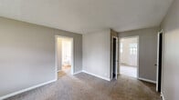 Located at 97 Patterson Village Drive in Dayton, Ohio, this single-floor unit offers three bedrooms, one bathroom, and a kitchen within a compact, efficiently arranged layout. The interior features neutral-toned walls throughout, paired with wall-to-wall carpeting in the living and bedroom areas that lends a consistent, understated feel across the space. Textured ceilings are present in several rooms, adding a subtle architectural detail. Each bedroom is equipped with windows fitted with horizontal blinds, allowing natural light to filter in while offering views of the surrounding greenery and exterior landscaping. Wall-mounted air conditioning units provide cooling in select rooms, while baseboard heaters run along the walls to provide warmth throughout the unit. The primary bedroom is generously sized relative to the overall footprint and includes direct access to the full bathroom. The bathroom features white tile surrounding a tub-shower combination, a window for natural light and ventilation, and clean, functional fixtures. The kitchen includes a full-size refrigerator and a window overlooking outdoor greenery, creating a bright workspace. A private entry door with natural light access is also present, completing the practical layout of this straightforward, move-in-ready unit.