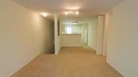 This single-floor space features an open layout connecting the living area to the kitchen. The living area is finished with neutral-toned carpet and is illuminated by a track lighting fixture, contributing to a bright and functional atmosphere. A ceiling fan with an integrated light is centrally positioned, providing both air circulation and additional lighting throughout the space.

The kitchen is partially separated from the living area by a raised pass-through counter, offering a convenient bar-style ledge while maintaining an open sightline. White appliances, including a refrigerator and what appears to be a washer or dryer unit, are visible within the kitchen area. A window above the counter allows natural light to filter into the kitchen, enhancing its brightness.

The layout is defined by a structural column and partial wall that divide the space without fully enclosing it, preserving the open feel. The neutral color palette of white walls and beige carpet creates a clean, understated aesthetic throughout. The space offers a practical and straightforward design suitable for everyday living.