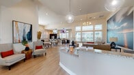 This well-appointed single-floor community clubhouse or leasing center is designed with a modern, welcoming aesthetic that blends functionality with sophisticated style. The space opens into a bright reception area featuring a granite-topped front desk, light hardwood flooring, and soaring vaulted ceilings that lend an airy, expansive feel throughout.

The main lounge area is a visual highlight, anchored by a dramatic large-scale photographic mural of a coastal pier at sunset. Comfortable seating arrangements, including plush sofas and accent chairs in neutral and warm orange tones, are layered over a bold geometric area rug, creating an inviting atmosphere. Floor-to-ceiling windows flood the area with natural light and offer outdoor views.

The kitchen and dining area is well-equipped with stainless steel appliances, dark cabinetry, granite countertops, and a spacious island bar with rattan bar stools. Wall-mounted televisions are thoughtfully positioned for viewing from multiple areas. A separate conference or meeting room features a large dark-wood table with seating, a wall-mounted display, and generous window access to natural light.

Throughout the space, carefully curated artwork and decorative elements add character, while the cohesive palette of whites, warm neutrals, and accent tones creates a polished, contemporary ambiance suitable for both professional and social gatherings.