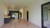 This single-floor space showcases a thoughtfully designed open-concept layout that seamlessly blends the kitchen and living areas into one cohesive environment. The kitchen is anchored by bold, dark cabinetry that creates a striking contrast against the light-toned walls and pale hardwood floors extending throughout the space. Stainless steel appliances, including a built-in wall oven, microwave, and a full-size refrigerator, lend a contemporary, professional-grade character to the kitchen. Recessed ceiling lighting provides even, modern illumination across the kitchen zone. A wall-mounted mini-split air conditioning unit ensures efficient climate control throughout the space. The open living area is bathed in a warm, neutral palette, with clean white walls and smooth ceilings that enhance the sense of openness and airiness. Dark interior doors add a sharp, modern accent and define the transitions between rooms. The layout offers a natural flow from the main living area toward additional rooms accessible via a central corridor, making the space both functional and easy to navigate. The combination of modern finishes, a neutral color scheme, and quality fixtures creates an inviting and practical environment well-suited for everyday living.
