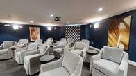 This single-floor screening room is thoughtfully designed to deliver a premium entertainment experience in an intimate setting. The space is defined by its deep navy blue walls, which create a dramatic, theater-like atmosphere while minimizing light reflection during screenings. A ceiling-mounted projector serves as the room's centerpiece technology, aimed toward a decorative geometric-patterned projection wall panel in black and white that doubles as a stylish focal point when not in use.

Seating consists of generously sized, plush light gray armchairs arranged in a loosely tiered layout, offering comfortable sightlines to the screen from multiple positions. Small round side tables are interspersed throughout the seating arrangement, providing convenient surfaces for guests. Wall-mounted sconces emit a warm, ambient glow, complementing the recessed ceiling lighting to create a balanced, moody illumination throughout the space.

Crown molding along the ceiling adds a refined architectural detail, elevating the overall aesthetic. The room's design successfully blends the functionality of a traditional screening room with the comfort and sophistication of an upscale lounge environment.