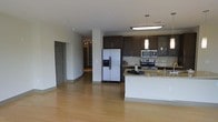 This single-floor residence offers a thoughtfully designed open-concept layout that seamlessly connects the kitchen and living areas, creating a spacious and airy feel. The contemporary kitchen is a standout feature, outfitted with rich dark cabinetry that provides striking contrast against the light granite countertops and a classic subway tile backsplash. Stainless steel appliances, including a side-by-side refrigerator with ice and water dispenser, a range, and an over-the-range microwave, lend a sleek, modern aesthetic. A central kitchen island with matching granite surfaces offers additional workspace and casual seating potential. Pendant lighting above the island adds a refined accent, complemented by recessed and flush-mount ceiling fixtures that ensure bright, even illumination throughout the space. Warm-toned hardwood flooring flows continuously across the open living area and into the hallway, tying the space together cohesively. Structural columns are subtly integrated into the layout, defining zones without interrupting the open flow. A corridor leads to additional rooms beyond, suggesting a well-organized floor plan. Overall, the space presents a clean, modern ambiance well-suited for contemporary living.