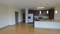 This single-floor residence offers a thoughtfully designed open-concept layout that seamlessly connects the kitchen and living areas, creating a spacious and airy feel. The contemporary kitchen is a standout feature, outfitted with rich dark cabinetry that provides striking contrast against the light granite countertops and a classic subway tile backsplash. Stainless steel appliances, including a side-by-side refrigerator with ice and water dispenser, a range, and an over-the-range microwave, lend a sleek, modern aesthetic. A central kitchen island with matching granite surfaces offers additional workspace and casual seating potential. Pendant lighting above the island adds a refined accent, complemented by recessed and flush-mount ceiling fixtures that ensure bright, even illumination throughout the space. Warm-toned hardwood flooring flows continuously across the open living area and into the hallway, tying the space together cohesively. Structural columns are subtly integrated into the layout, defining zones without interrupting the open flow. A corridor leads to additional rooms beyond, suggesting a well-organized floor plan. Overall, the space presents a clean, modern ambiance well-suited for contemporary living.