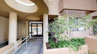 This building makes a strong first impression with its warm, earth-toned stucco exterior and expressive architectural design. The façade is characterized by curved archways, rounded overhangs, and layered geometric forms that lend the structure a distinctive, sculptural quality. A welcoming entrance is framed by raised planter beds filled with lush greenery, small trees, and seasonal flowering plants, adding vibrant color to the approach. The entryway is fully accessible, featuring a gently sloping ramp with metal handrails leading to a set of glass double doors. Above the entrance, a striking oval skylight cutout allows natural light to filter into the covered vestibule, enhancing the sense of openness. Glass block panels along the entry corridor further diffuse light while maintaining privacy. Flagpoles flank the main approach, and a tree-lined sidewalk borders the property, softening the urban streetscape. The building's multi-story elevation includes balconies and large windows that suggest well-lit interior spaces throughout. The overall ambiance is warm, inviting, and thoughtfully designed, blending functional accessibility with architectural character that creates a welcoming environment for all who enter.