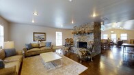 This single-floor space offers a welcoming and well-appointed layout designed for both comfort and gathering. The living area serves as the heart of the space, anchored by a striking floor-to-ceiling stone fireplace that becomes an immediate focal point. Crafted from natural river rock, the fireplace lends a rustic, timeless character to the room. The living area features warm-toned hardwood flooring partially covered by a soft area rug, with recessed lighting overhead illuminating the space. Comfortable seating, including a leather sofa and ornate upholstered accent chairs, is arranged to encourage conversation and relaxation. A framed landscape painting adorns one wall, adding a refined artistic touch. Natural light filters in through nearby windows, further brightening the interior. Adjacent to the living area, a dining space is visible, offering additional room for entertaining. The space also connects to an impressive outdoor amenity: a covered patio area with a vaulted wood-paneled ceiling and ceiling fan, which opens to a fenced pool. The poolside patio is furnished with wrought iron tables and lounge chairs, providing a shaded retreat ideal for outdoor leisure, set against a backdrop of lush mature trees.