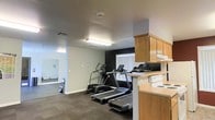 This single-floor fitness and amenity space is designed to support an active lifestyle in a practical, no-frills layout. The workout area features dark carpeted flooring that provides cushioning underfoot and houses a selection of cardio equipment, including two treadmills and an elliptical trainer. A dark accent wall at the back of the room adds a modern contrast to the otherwise light-toned walls, while a window with vertical blinds allows natural light to filter in, brightening the space. Overhead fluorescent lighting ensures consistent illumination throughout. Adjacent to the fitness area is a secondary zone featuring warm-toned wooden cabinetry and a countertop, suggesting a utility or lounge area that complements the workout space. A mirror is mounted on the far wall of the fitness area, adding depth and functionality. The ceiling is finished with a standard texture, and HVAC vents are integrated to maintain airflow. Overall, the space offers a straightforward, utilitarian environment well-suited for daily exercise and light amenity use.