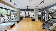 Located at 1125 S Second St in Minneapolis, MN, this fitness center offers a generously proportioned, single-floor exercise space designed with both functionality and aesthetics in mind. The room features warm hardwood-style flooring throughout, providing a clean and durable surface suited for a wide range of workout activities.

The cardio zone is well-appointed with multiple treadmills, stationary bikes, and a StairMaster, all arranged to allow comfortable spacing between users. A dedicated strength training area includes a comprehensive dumbbell rack with weights of varying sizes, cable machines, and a squat/power rack, catering to a full range of resistance training needs. Fitness accessories such as medicine balls, resistance bands, and plyo boxes are also available for functional and cross-training workouts.

Concrete columns accent the space, contributing to a modern industrial aesthetic that is complemented by a bold graphic mural on the feature wall. Track lighting runs along the white ceiling, ensuring the space is brightly and evenly lit. Ceiling fans provide additional airflow and comfort during workouts. A wall-mounted flat-screen television adds an entertainment element to the space.

Large windows along one side of the room invite natural light and provide views to the outdoors, while mirrors strategically placed throughout the space enhance its openness. The overall ambiance is contemporary, clean, and purpose-built for an effective fitness experience.