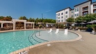 Located at 4001 Reedybrook Crossing in Apex, NC, this outdoor amenity space is designed to deliver a resort-style experience within a residential community setting. At the heart of the area is a generously sized, freeform swimming pool with a shallow sun shelf featuring in-water lounge chairs — ideal for relaxing under the North Carolina sky. The pool deck is lined with chaise lounges and market umbrellas, providing ample space for sunbathing and leisure. A classic white pergola with string lights anchors one corner of the deck and shelters a built-in outdoor kitchen with a stone surround, perfect for al fresco dining and entertaining. The space is accessed through a gated entry with clearly posted pool rules, ensuring a secure and organized environment. Well-maintained landscaping throughout — including ornamental grasses, flowering shrubs, mulched beds, and mature trees — frames the amenity area with natural beauty and visual interest. Concrete walkways connect the pool area to the surrounding buildings, which feature a mix of horizontal siding, stone accents, and large glass panels. The overall ambiance is bright, open, and inviting, with scenic views of the lush tree line beyond the property perimeter completing the setting.