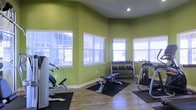 This single-floor fitness space is designed to offer a functional and energizing workout environment. The room is finished with bold, lime-green walls that create a lively and motivating atmosphere, complemented by crisp white baseboards and trim. Natural light flows generously into the space through multiple large windows fitted with white horizontal blinds, allowing users to control light levels while maintaining a bright, open feel.

The flooring consists of light-toned hardwood-style planks, adding warmth and a clean aesthetic to the room. Recessed lighting in the ceiling provides additional illumination. The layout is thoughtfully organized to accommodate a variety of workout styles. A multi-tier dumbbell rack stocked with a range of free weights is positioned along one wall, paired with an adjustable workout bench centered on a protective rubber mat. Strength training machines are positioned toward the corners of the room, making efficient use of the available floor space. The angled ceiling adds a subtle architectural interest and contributes to the room's sense of openness. Overall, the space is clean, well-lit, and equipped to support a comprehensive fitness routine.