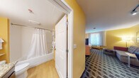This single-floor hotel suite is designed to offer the comfort and functionality of a home away from home. The space is characterized by warm golden-yellow walls, patterned geometric carpeting throughout, and light wood accents that create a welcoming and cohesive aesthetic.

The sleeping area features a queen bed dressed in crisp white linens, a flat-screen TV mounted on a wood media console, and a bedside lamp that adds a warm glow. A window with sheer curtains allows natural light to fill the room, contributing to an airy atmosphere.

The open living area includes a sofa, an armchair, and a dedicated workspace equipped with a built-in desk, overhead shelving, and an ergonomic chair — well-suited for productivity. A flat-screen TV is also accessible from this area.

The kitchenette is thoughtfully outfitted with dark cabinetry, granite-style countertops, a stainless steel sink, a microwave, and a stove, providing ample resources for in-suite meal preparation.

The bathroom features light hardwood-style flooring, a granite-topped vanity with dark wood cabinetry, a full bathtub with a shower curtain, and grab bars for added accessibility. The suite's layout flows seamlessly from one functional zone to the next, making it an efficient and comfortable space for extended stays.
