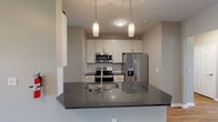 This single-floor residence offers a well-designed open-concept layout with a neutral gray color palette and warm wood-style flooring that flows through the main living areas. The kitchen serves as a central feature, showcasing dark granite countertops on both the peninsula and surrounding surfaces, crisp white shaker-style cabinetry extending to the ceiling, and a full suite of stainless steel appliances including a French door refrigerator, electric range, and built-in microwave. Dual pendant lights hang above the peninsula, providing both task and ambient lighting, while the open pass-through connects seamlessly to the spacious living area. The living space is generously proportioned, with multiple doorways leading to the bedrooms and additional rooms. A dedicated utility corridor beside the kitchen offers additional counter and cabinet space with a classic subway tile backsplash. The bathrooms include a full tub-shower combination with a curved curtain rod and a separate primary bathroom featuring a tiled walk-in shower with glass doors, a vanity with matching granite countertops, and direct access to a walk-in closet. Bedroom spaces are carpeted and receive natural light through large windows with exterior views.