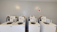 Located at 20 Old Yellow Springs Road in Fairborn, Ohio, this single-floor shared laundry facility is designed to serve the practical needs of its residents. The space is clean and functional, with neutral gray walls that contribute to a bright, orderly environment. The room houses a row of coin-operated, top-load washing machines lined up side by side along the wall, each clearly labeled and equipped with easy-to-read operating instructions. The machines are maintained in white finish, giving the space a tidy, uniform appearance. Signage is posted on the wall to provide users with guidelines and important notices regarding proper use of the equipment. The layout is straightforward and efficient, allowing multiple users to operate machines simultaneously with ease. The flooring is practical and durable, suited to the demands of a high-traffic laundry environment. Overall, this laundry facility provides a convenient, no-frills amenity that prioritizes functionality and accessibility for all users of the property.