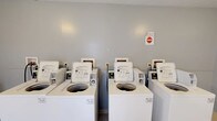 Located at 20 Old Yellow Springs Road in Fairborn, Ohio, this single-floor shared laundry facility is designed to serve the practical needs of its residents. The space is clean and functional, with neutral gray walls that contribute to a bright, orderly environment. The room houses a row of coin-operated, top-load washing machines lined up side by side along the wall, each clearly labeled and equipped with easy-to-read operating instructions. The machines are maintained in white finish, giving the space a tidy, uniform appearance. Signage is posted on the wall to provide users with guidelines and important notices regarding proper use of the equipment. The layout is straightforward and efficient, allowing multiple users to operate machines simultaneously with ease. The flooring is practical and durable, suited to the demands of a high-traffic laundry environment. Overall, this laundry facility provides a convenient, no-frills amenity that prioritizes functionality and accessibility for all users of the property.