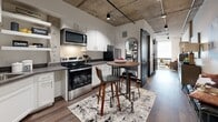 Located at 708 22nd Street in Greeley, Colorado, this single-floor unit delivers a striking industrial-modern aesthetic within an efficiently designed footprint. Exposed concrete ceilings with visible structural elements and track lighting set a bold architectural tone throughout the space, complemented by warm hardwood-style flooring that runs continuously across the main areas.

The kitchen is a standout feature, outfitted with white shaker cabinetry, dark granite-style countertops, and a full suite of stainless steel appliances including a range, microwave, dishwasher, and refrigerator. A compact dining nook with a bistro-style table and bar seating integrates seamlessly into the kitchen area, making it both functional and social.

The living area extends from the kitchen in an open-plan arrangement, offering a comfortable and versatile space anchored by industrial-chic design details. Exposed concrete columns punctuate the room, adding architectural character and reinforcing the urban loft atmosphere.

The bathroom is clean and contemporary, featuring wood-toned flooring, a wall-mounted vanity with open storage, a mirrored medicine cabinet, and crisp white walls. Black matte fixtures and trim details tie the bathroom's design back to the broader aesthetic of the unit.

Overall, this space balances compact, efficient living with a distinctive urban character rooted in raw materials and modern finishes.