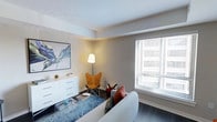 This thoughtfully designed single-floor space showcases a clean, contemporary aesthetic that balances comfort with modern style. The living area serves as the heart of the space, featuring dark hardwood flooring that contrasts beautifully against the crisp white walls and ceiling. A large window fitted with white horizontal blinds allows an abundance of natural light to fill the room, creating a bright and airy ambiance. The space is elegantly furnished with a plush light grey sofa accented by vibrant red and patterned throw pillows, complemented by a sleek acrylic coffee table atop a rich blue area rug. A distinctive amber butterfly chair and a coordinating floor lamp add a warm, sculptural focal point to the corner of the room. A low-profile white dresser anchors one wall, topped with decorative accents, while a bold landscape artwork adds a pop of color and artistic character to the space. Crown-style ceiling trim details lend a refined architectural touch throughout. The overall ambiance is modern yet welcoming, with a carefully curated mix of textures, colors, and materials that come together to create a stylish and functional living environment.