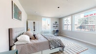 This thoughtfully designed studio unit offers a bright, open-concept layout spread across a single floor, blending living, sleeping, and cooking areas seamlessly. The sleeping area is generously proportioned, featuring light hardwood-style flooring, neutral-toned walls, and multiple large windows that flood the space with natural light while offering views of the surrounding urban landscape. A white wardrobe cabinet provides ample storage, complemented by an open shelving unit ideal for a home office or study nook.

The kitchen is efficiently designed along a single wall and features gray cabinetry with sleek hardware, white subway tile backsplash, light countertops, a stainless steel sink with a modern gooseneck faucet, and a full suite of appliances including a range, microwave, and refrigerator. Open shelving above the counter adds both display space and accessibility.

The bathroom maintains the unit's clean, modern aesthetic with large-format wood-look tile flooring, a wall-mounted vanity with a wide mirror, and a window that brings in natural light. The overall ambiance of the space is contemporary and airy, with a neutral color palette and consistent use of light materials throughout that create a cohesive, welcoming feel.
