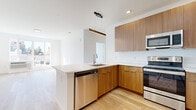 Located at 22 W Camden in Hackensack, NJ, this single-floor unit showcases a contemporary design characterized by clean lines, bright white walls, and light hardwood flooring that flows consistently throughout the space. The open-concept layout seamlessly connects the kitchen and living areas, creating a cohesive and functional environment.

The kitchen is a standout feature, equipped with flat-front cabinetry in a warm wood tone, striking veined quartz countertops, and a peninsula with an undermount sink. Stainless steel appliances, including a range, microwave, and dishwasher, complement the modern aesthetic. Pendant lighting above the peninsula adds a refined touch.

The living area is bathed in natural light from large windows that frame views of the surrounding Hackensack skyline, enhancing the open and airy atmosphere. The bedroom spaces are clean and minimalist, finished with the same hardwood flooring and crisp white walls, offering a calm and neutral backdrop.

The bathroom continues the modern theme with large-format tile surrounding the soaking tub and shower combination, a wood-toned vanity with a white countertop, and sleek chrome fixtures, completing the unit's cohesive and well-appointed design.
