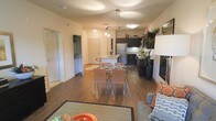 This single-floor residence showcases an open-concept design that seamlessly connects the living, dining, and kitchen areas into one cohesive and functional space. Rich hardwood-style flooring extends throughout, adding warmth and continuity across the main living areas.

The kitchen is equipped with dark cabinetry contrasting against light countertops, stainless steel appliances including a full-size refrigerator, and a peninsula island that provides additional workspace and casual seating. A pendant light fixture with multiple hanging lights illuminates the kitchen and dining zone, enhancing the modern aesthetic.

The dining area features a glass-top table paired with contemporary chairs, positioned centrally to take advantage of the open layout. A textured area rug anchors the living space, which includes a comfortable sectional with accent pillows in coordinating tones.

Tall indoor plants are strategically placed throughout, introducing natural elements to the interior. A hallway visible from the main area leads to additional rooms, suggesting a practical and well-organized floor plan. The overall ambiance is modern, clean, and inviting, with warm neutral tones throughout.