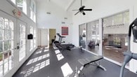 This fitness center offers a bright, open workout environment designed to support a well-rounded exercise routine. The space is defined by dramatically high ceilings that create an airy, open atmosphere, while crisp white walls and large windows allow abundant natural light to flood the interior throughout the day. Rubber flooring covers the entire workout area, providing both safety and durability underfoot.

The facility is outfitted with a comprehensive selection of cardio equipment, including an elliptical trainer, a rowing machine, and a treadmill, allowing users to vary their cardiovascular workouts. Strength and functional training is supported by an adjustable weight bench and balance equipment such as BOSU balls. A ceiling fan circulates air throughout the space, enhancing comfort during workouts.

Elegant French doors with glass panes serve as a divider between the fitness area and an adjoining room that appears to function as an office or business center, complete with workstations and seating. Large sliding glass doors open toward an outdoor area, further connecting the interior to the surrounding environment. The overall design balances functionality with a clean, modern aesthetic, making this fitness center a practical and inviting space for daily exercise.