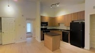 Located at 2904 Calvary Dr in Raleigh, North Carolina, this single-floor residence offers a thoughtfully designed layout with a warm and welcoming ambiance. The kitchen serves as a central highlight of the space, showcasing honey-toned wood cabinetry that extends to impressive heights, taking full advantage of the soaring ceilings. A center island with a dark countertop provides additional workspace and a natural gathering point. Black appliances, including a range, microwave, dishwasher, and refrigerator, offer a sleek contrast to the cabinetry, while track lighting overhead ensures the area is well-lit. Light hardwood flooring runs throughout, lending a clean, cohesive feel to the interior and reflecting natural light effectively. A hallway visible from the kitchen leads to additional areas of the home, with a glimpse of natural light streaming through a door or window at the far end, hinting at the brightness carried throughout the residence. The tall ceilings create an open, airy atmosphere, adding a sense of spaciousness to the overall design. The combination of warm wood tones, dark countertops, and light flooring gives this space a balanced and functional aesthetic well-suited for everyday living.