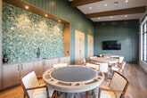 The Culprr Room is a thoughtfully designed gaming and social lounge located at 303 Arrowhead Ln in South Setauket, NY. The single-floor space is anchored by multiple dedicated poker and card tables, each surrounded by comfortable mid-century-modern inspired wooden chairs with upholstered cushions, creating an inviting setting for both casual and competitive play.

The room's design strikes a balance between sophisticated and relaxed. Vertical shiplap-style paneling in a muted sage green lines the walls, complemented by warm wood tones throughout, including light hardwood flooring and exposed wood beam accents along the ceiling. A standout feature is the built-in wet bar area, framed by a dramatic botanical-patterned accent wall with recessed lighting, flanked by floor-to-ceiling cabinetry in a natural wood finish — ideal for refreshments during gatherings.

Large, multi-pane steel-framed windows along one wall flood the space with abundant natural light while offering views of the outdoor surroundings. A wall-mounted flat-screen television adds a media element to the room. A sleek glass partition wall with black steel framing separates the lounge from an adjacent common area, maintaining visual connectivity while defining the space. The overall ambiance is modern, warm, and purposefully curated for social entertainment.