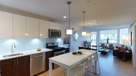 Located at 145 Westerly Rd in Ossining, New York, this single-floor residence offers a thoughtfully designed open-concept layout that seamlessly connects its living, dining, and kitchen areas. The modern kitchen is a standout feature, equipped with stainless steel appliances including a gas range, built-in microwave, and dishwasher. Sleek white cabinetry with under-cabinet lighting lines the walls, complemented by a light blue glass tile backsplash that adds a subtle pop of color. Recessed ceiling lights and stylish pendant fixtures illuminate the space with a clean, contemporary feel. A white dining table with modern chairs sits at the center of the eat-in area, perfectly positioned beneath pendant lighting and adjacent to a window that draws in natural light. The open flow leads into multiple living areas furnished with comfortable seating arrangements and accented by natural light from several windows throughout. The floor plan reveals a well-proportioned layout with distinct zones for relaxing, dining, and entertaining, all maintaining a cohesive and airy ambiance. The neutral color palette and modern design choices throughout contribute to a bright, welcoming atmosphere suited for everyday living.